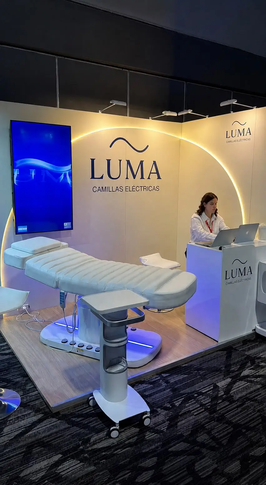 Stand de LUMA en un congreso médico: camilla eléctrica Premium blanca con iluminación LED en la base, carro auxiliar, pantalla de marca y personal atendiendo en el mostrador.